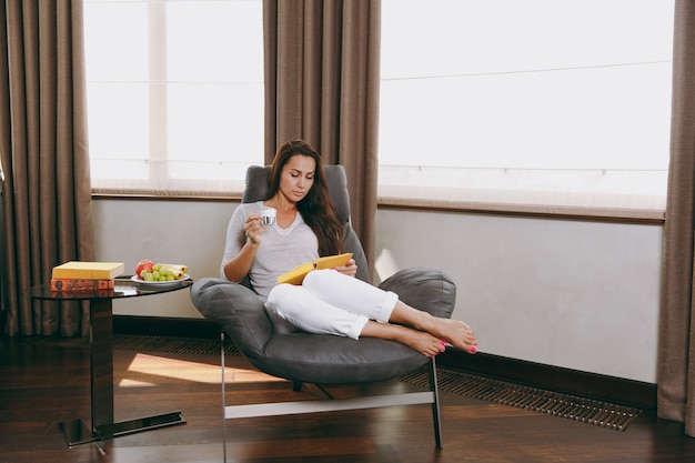 写真 家の美しい若い女性は、窓の前のモダンな椅子に座って、リビングルームでリラックスし、本を読み、コーヒーやお茶を飲みます。
