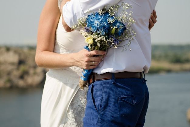 Фото Невеста обнимает жениха и держит букет невесты с голубыми цветами на фоне реки.