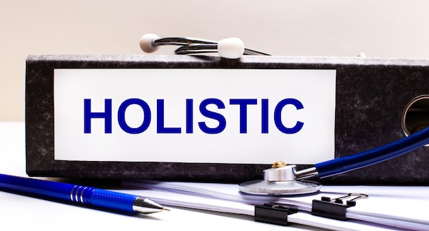 На рабочем столе есть стетоскоп, синяя ручка и серая папка с текстом holistic medical concept.