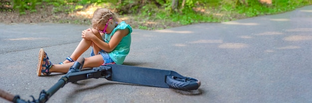 写真 少女は公園でスクーターに乗っているときに転倒し、ひざをひどく傷つけた。電動スクーター傷害の概念。テキストの場所を含むパノラマ写真