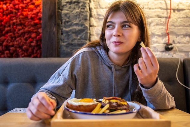 写真 カフェの女の子は大きなハンバーガーとフライドポテトを楽しんでいます。