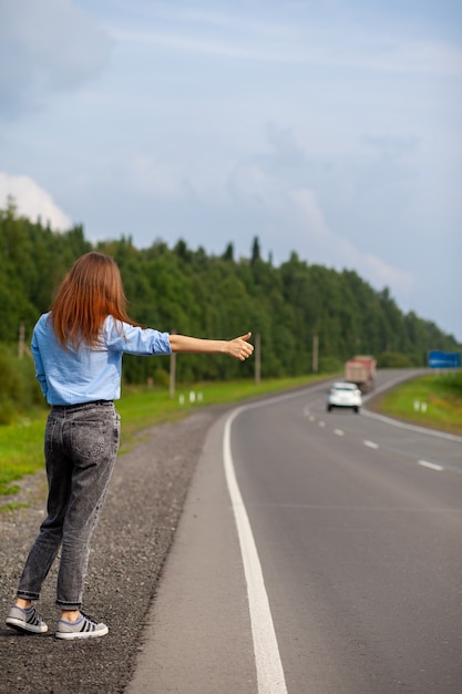 Фото Девушка рукой останавливает машину на трассе. стильная женщина на дороге останавливает машину, отправляется в путешествие. дорога посреди леса.