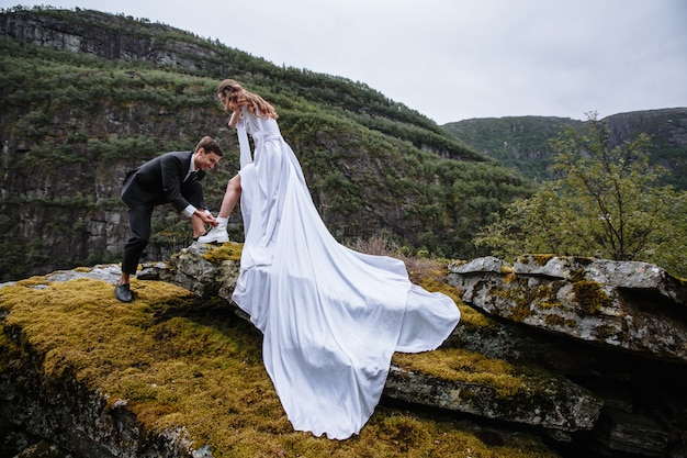Фото Жених помогает невесте, завязывая шнурок на ботинке. портрет пары свадьбы стоя на утесе.