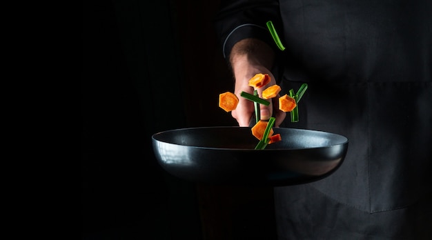 Фото Рука профессионального повара бросает кусочки овощей на сковороду на черном фоне. концепция приготовления пищи в ресторане. бесплатное рекламное место