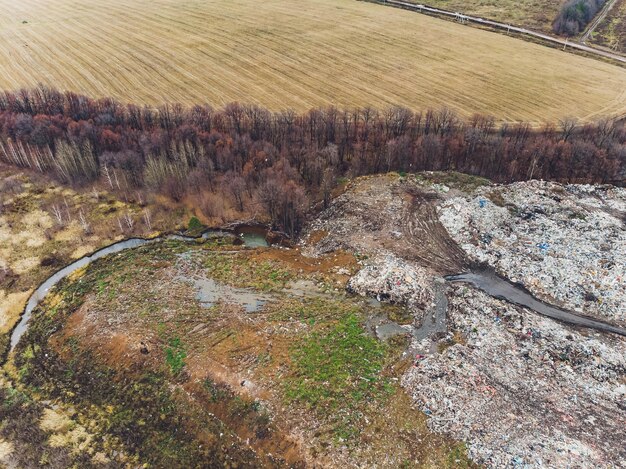 Фото Большая куча мусорной свалки простирается параллельно реке. горы мусора с облачным небом на фоне земли в дневное время. отходы нефтепродукты являются основными.