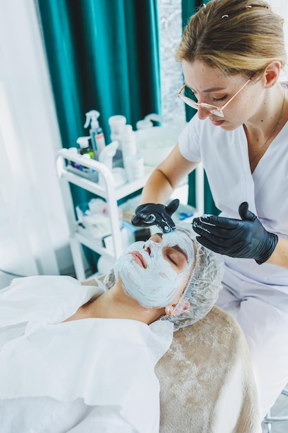 Фото Офис косметолога, наносящего маску на лицо женщины в спа-салоне, вид над эстетической процедурой маска по уходу за кожей лица