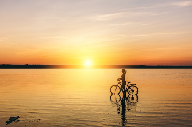 暖かい夏の日の日没時に水中で自転車に座っているスーツを着たスポーティな女の子のシルエット。フィットネスのコンセプト。