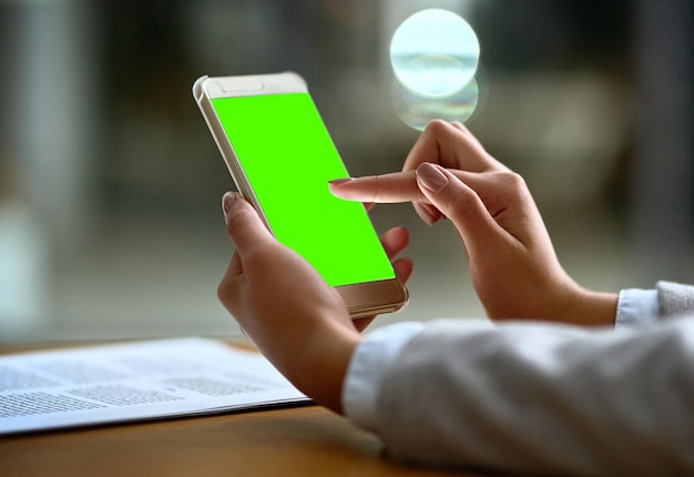 写真 仕事の日を整理する最も賢明な方法 - ビジネスウーマンがデスクに緑色の画面の携帯電話を使って - 現代のオフィスパーソン