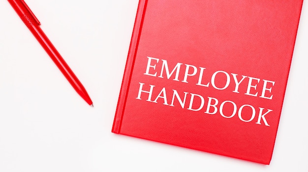 Employee handbookというテキストは、オフィスの白いテーブルの赤いペンの近くにある赤いメモ帳に書かれています。ビジネスコンセプト