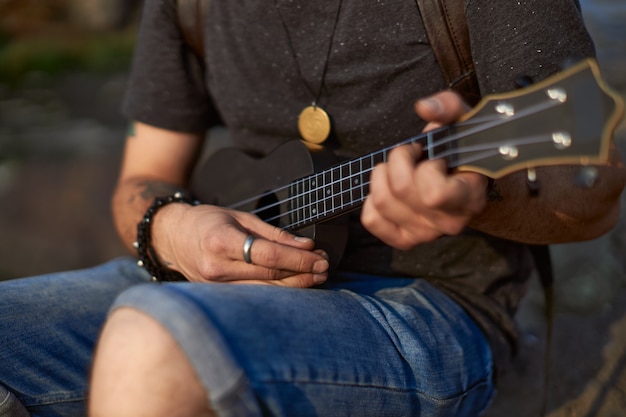 Фото Руки молодого человека играют на черной гавайской гитаре, сидя на камне, концепция творчества высокого качества ...