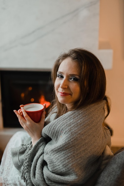 Фото Молодая женщина, покрытая уютным теплым одеялом, держит чашку горячего напитка женщина сидит с закрытыми глазами на фоне камина