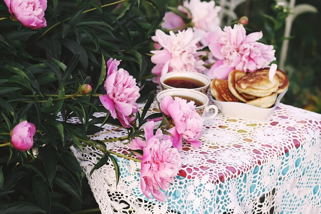 Thee in landelijke stijl in de zomertuin in het dorp Twee kopjes zwarte thee en handgemaakte pannenkoeken op gehaakt vintage kanten tafelkleed en bloeiende pioenrozen in zonlicht
