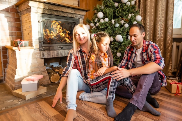 テーマクリスマスと新年の家族サークル。暖炉のクリスマスツリーの近くのリビングルームの家の木の床に座っている若い白人家族。