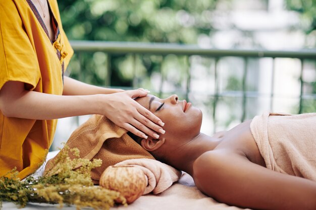 Foto terapista che massaggia la testa e il viso della giovane e bella donna afro-americana rilassante in località termale