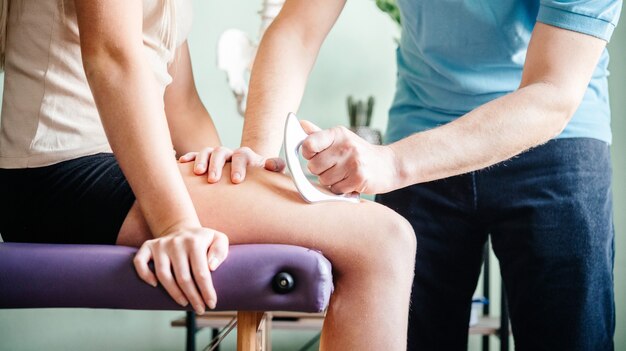 Therapist using IASTM instrument treatment, girl receiving soft tissue treatment on her leg with guasha stainless steel tool