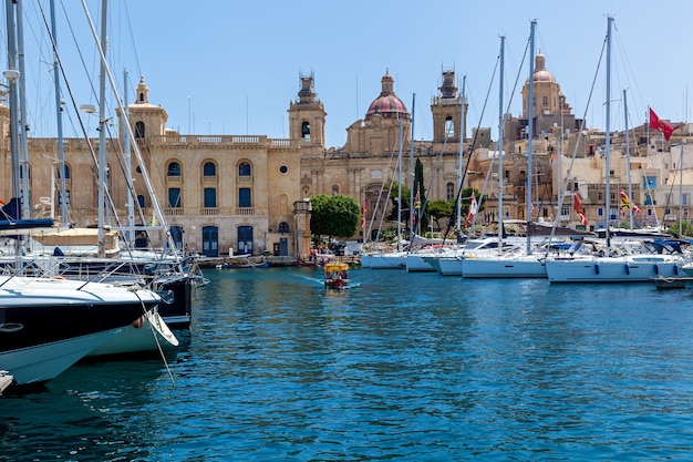 There are a lot of moored yachts in the harbor on the background of Maltese city with old buildings and palm trees on a summer sunny day.