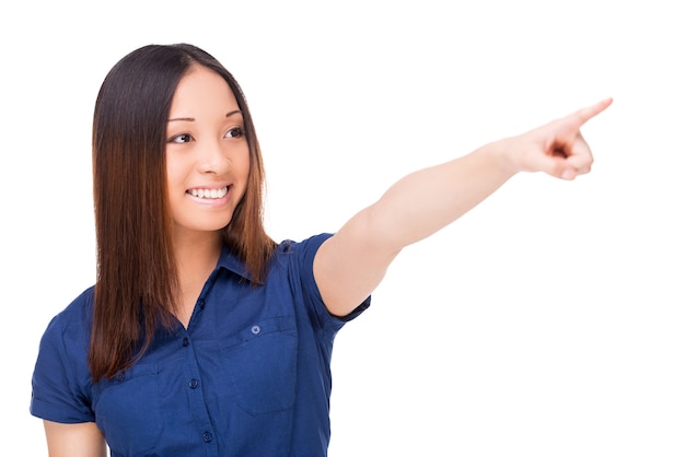 Over there! Beautiful young Asian woman pointing away and smiling while standing isolated on white