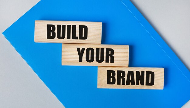 There is a blue notebook on a light gray background. Above are three wooden blocks with the words BUILD YOUR BRAND.