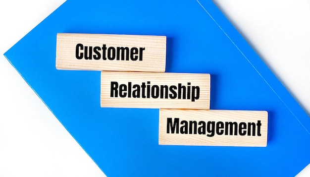 明るい灰色の背景に青いノートがあります 上にあるのは CRM Customer Relationship Management という言葉が書かれた 3 つの木製のブロックです