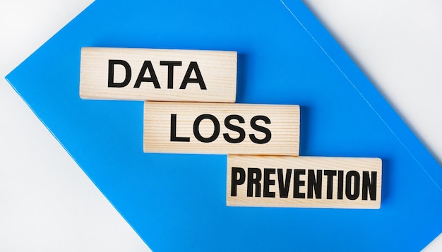 밝은 회색 배경에 파란색 노트북이 있습니다. 위는 DATA LOSS PREVENTION이라는 단어가 있는 세 개의 나무 블록입니다.