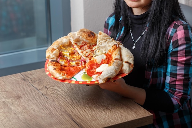 There is a box with different types of pizzas and shawarma on the table, women's hands are holding,