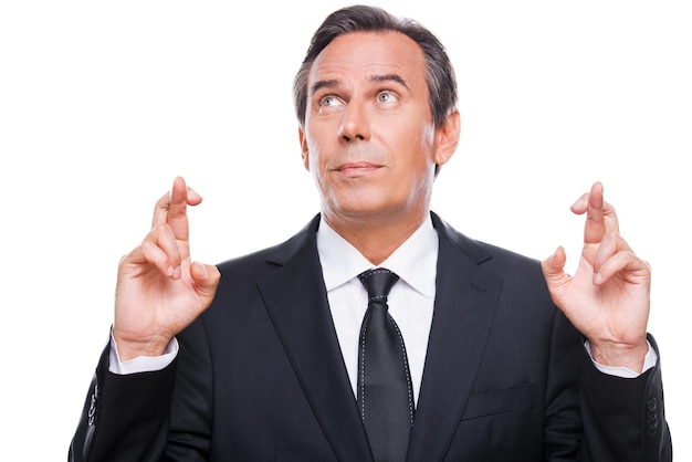 There is a hope... Portrait of mature man in formalwear keeping fingers crossed and eyes closed while standing isolated on white background