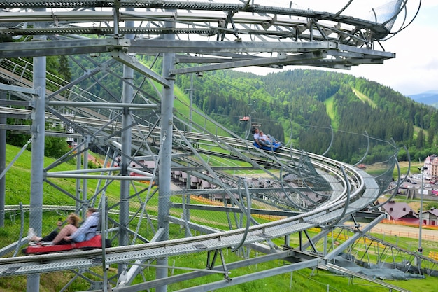 There is a roller coaster ride on the summer slope of the mountain. Two booths with people descend along it, against a blurred background. People are impersonal