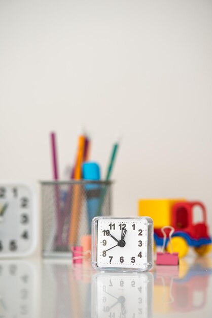 C'è un orologio quadrato bianco in primo piano su uno sfondo di cancelleria sfocata e giocattoli per bambini