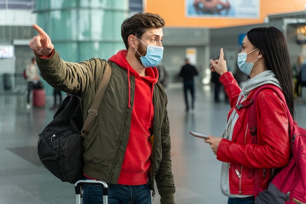 There, no there. Waist up portrait view of the couple wearing protective masks argue with each other while pointing at the different ways at the airport. Traveling during quarantine concept