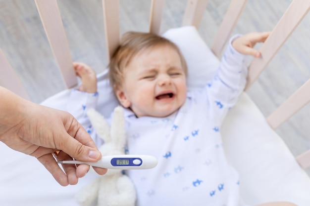 Thermometer in focus in moeders handen met een hoge temperatuur van 39 graden op de achtergrond van een zieke huilende babyjongen in een wieg thuis op wit beddengoed, geneeskunde en kindergeneeskunde