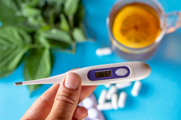 A thermometer showing a temperature of 385 degrees in the hand against the background of tea with lemon and tablets medicines The concept of seasonal diseases the treatment of diseases