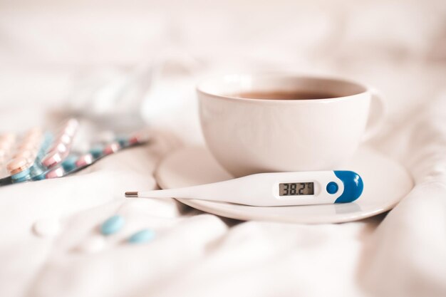 Thermometer with high temperature on plate and cup of tea with pills closeup in bed. Good morning. Illness concept. Social isolation.