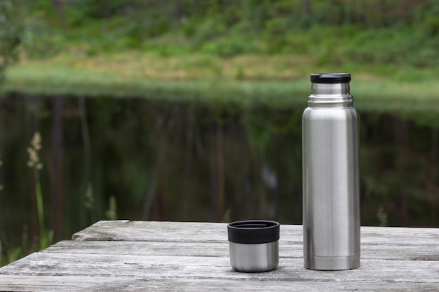 Photo thermos and cup on wooden table in forest near lake