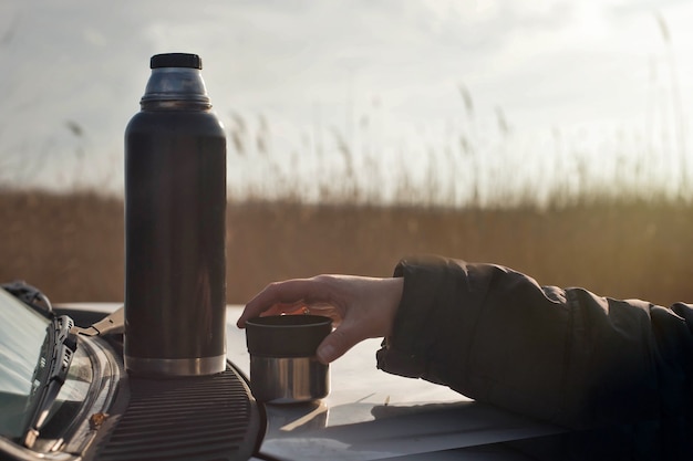 Thermos and hand with cup of tea on the hood of the car. Rest and relax.