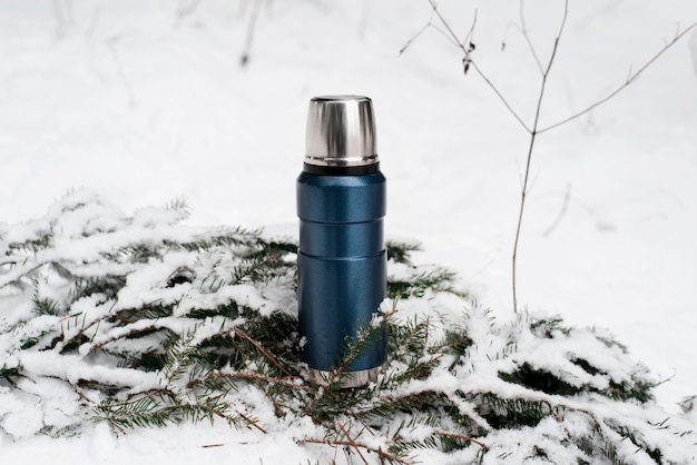Thermos staande op dennentak in de sneeuw. Vacuümfles buitenshuis. Winter kamperen in het bos-concept.
