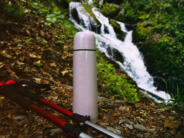 Thermos and trekking poles at defocused natural background with waterfall