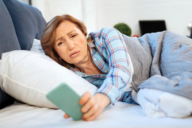 Under thick blanket. Upset mature woman taking smartphone from nightstand and checking time on screen while lying in bed