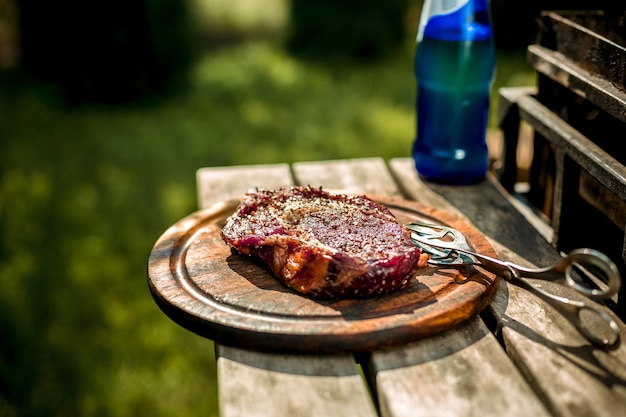 A thick strip steak being grilled outdoors. A large piece of meat on a wooden board next to the grill. Cooking food