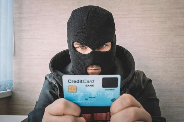 Вор в черной маске держит в руках украденную банковскую карту. Квалифицированный преступник в маске использует украденную кредитную карту для покупки вещей в Интернете.