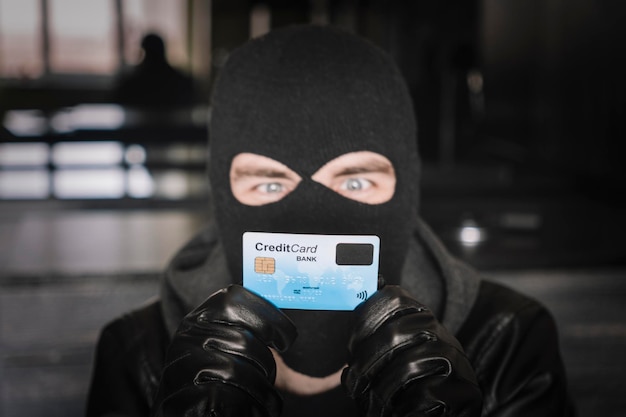 Foto un ladro con una maschera nera tiene in mano una carta di credito rubata abile criminale mascherato che utilizza una carta di credito rubata per acquistare oggetti online