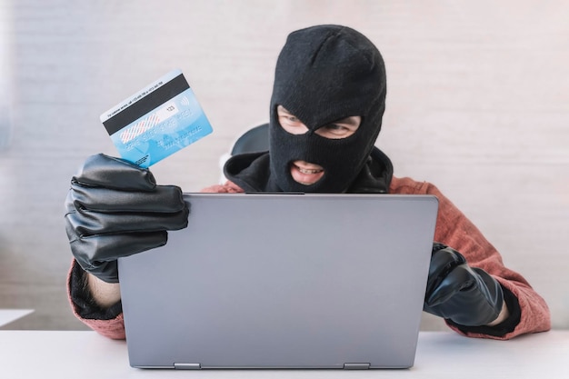 Thieves hold credit cards using laptop computer for password hacking activities Cyber crime concepts a male thief holds a credit card in his hand and picks up a password to steal money