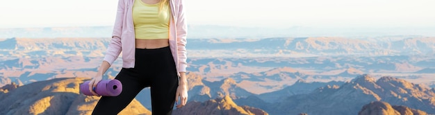 A thin athletic girl takes a break between classes on the background of mountains in the early morning