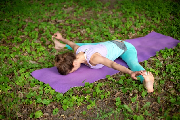 Thin brunette girl plays sports and performs beautiful and sophisticated yoga poses in a summer park.