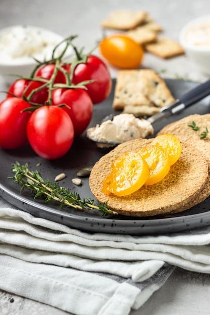 Thin crispy wholegrain crackers with cream cheese and tomatoes on a black plate. 