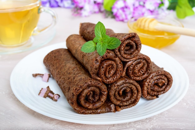 Thin delicate chocolate pancakes, rolled, laid out in a pile on a white plate and a cup of herbal tea with honey