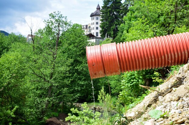 A thin stream of water flows from a sewer plastic pipe against a blurred background of trees