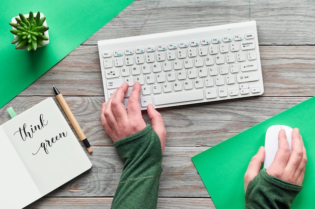 Think green, zero waste creative flat lay. Top view with hands typing on keyboard and holding computer mouse. Open notebook with motivation text, minimal wooden workspace with succulent plant on wood.