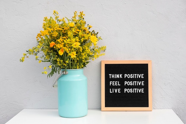 긍정적인 느낌을 긍정적으로 생각하고 편지판에 긍정적인 동기 부여 인용문을 쓰고 회색 돌담에 대고 흰색 테이블에 노란색 꽃을 부케합니다. 오늘의 영감을 주는 인용문