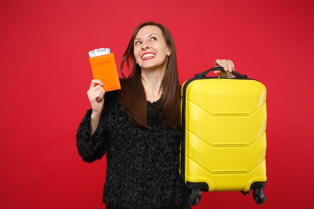 Thinkful young woman in black fur sweater looking up, holding suitcase, passport boarding pass ticket isolated on bright red background. People sincere emotions, lifestyle concept. Mock up copy space.