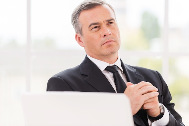 Foto pensando agli affari. uomo maturo premuroso in abiti da cerimonia che tiene le mani giunte e distoglie lo sguardo mentre è seduto al suo posto di lavoro